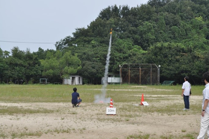 高専スペース連携での活動のようす。広い空き地で上空に向かって飛ぶ小型ロケットの行方を見守る人々。