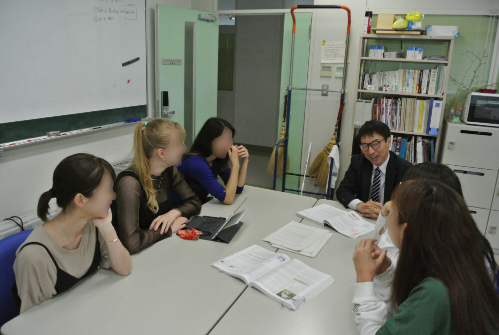 テーブルを囲んで芥川先生と女子学生5名と議論している場面。留学生も。