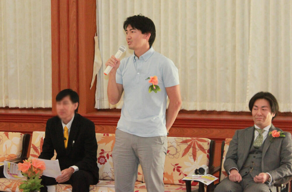 薄い水色のポロシャツに、胸元に華やかなオレンジ色のお花を挿してスピーチをする前澤先生。