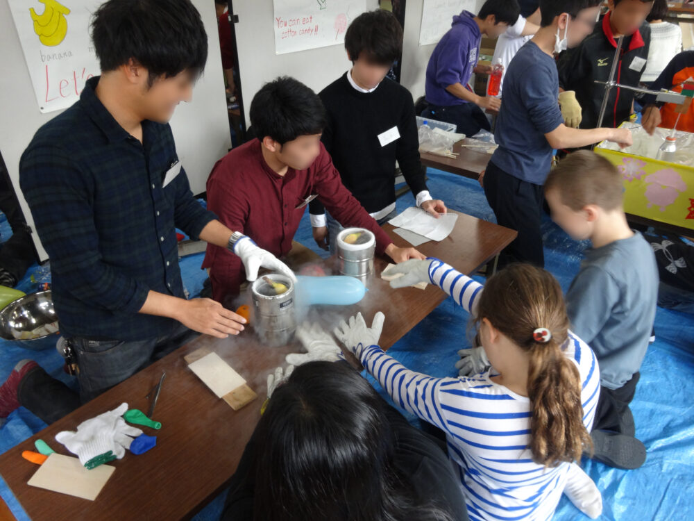 外国の子どもと日本の学生がひとつのテーブルで実験している。真ん中の実験器具からは白い霧のようなものが発生している。