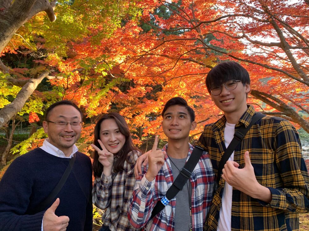 赤・オレンジ・黄・黄緑・緑、鮮やかな紅葉の木々を背景に、留学生3名とともに写真におさまる畑村先生。