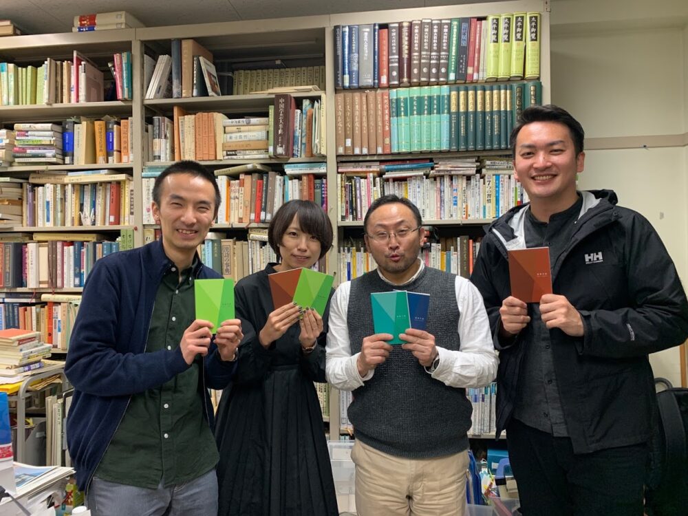 畑村先生の研究室で、高専手帳を手に集合写真を撮る畑村先生。浅村さん・笹尾さんらとともに。