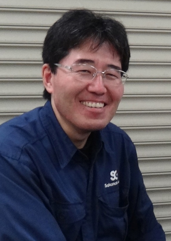 山本さんの上半身の写真。紺色の作業着をきて、柔らかくはにかんでいる。