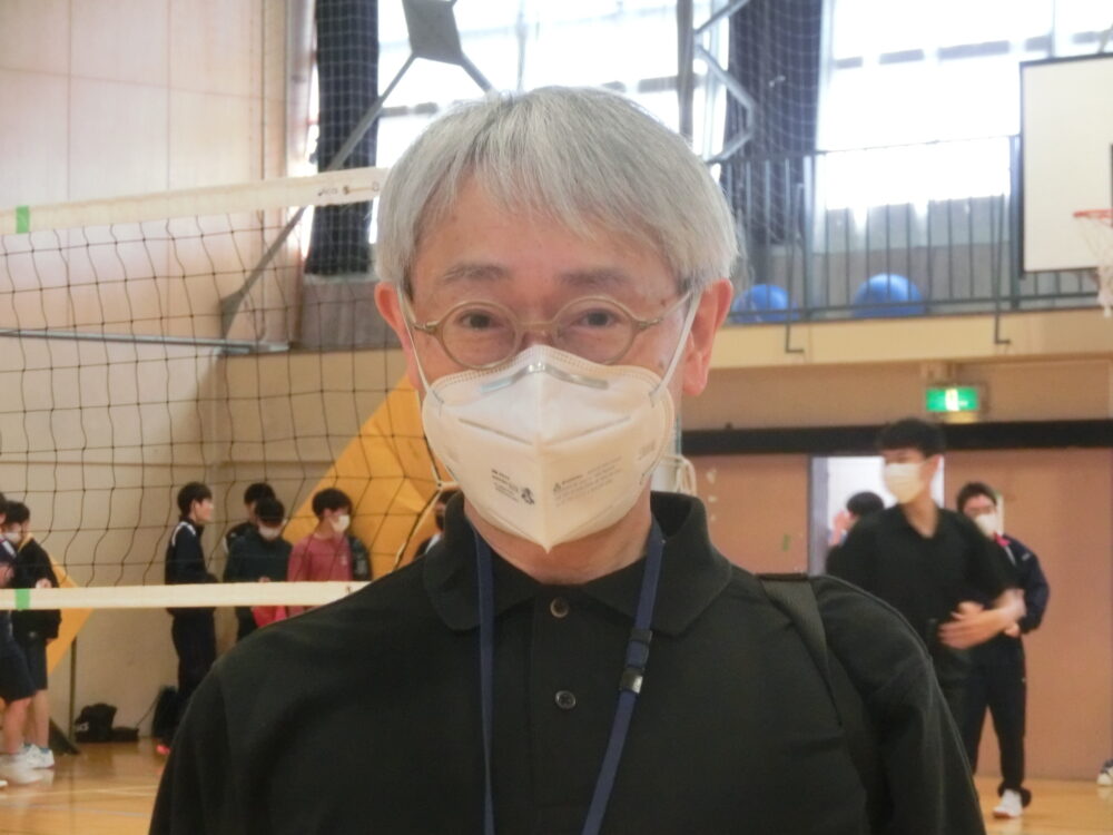 体育館のバレーボールネットを背景に、松江先生の上半身のお写真。