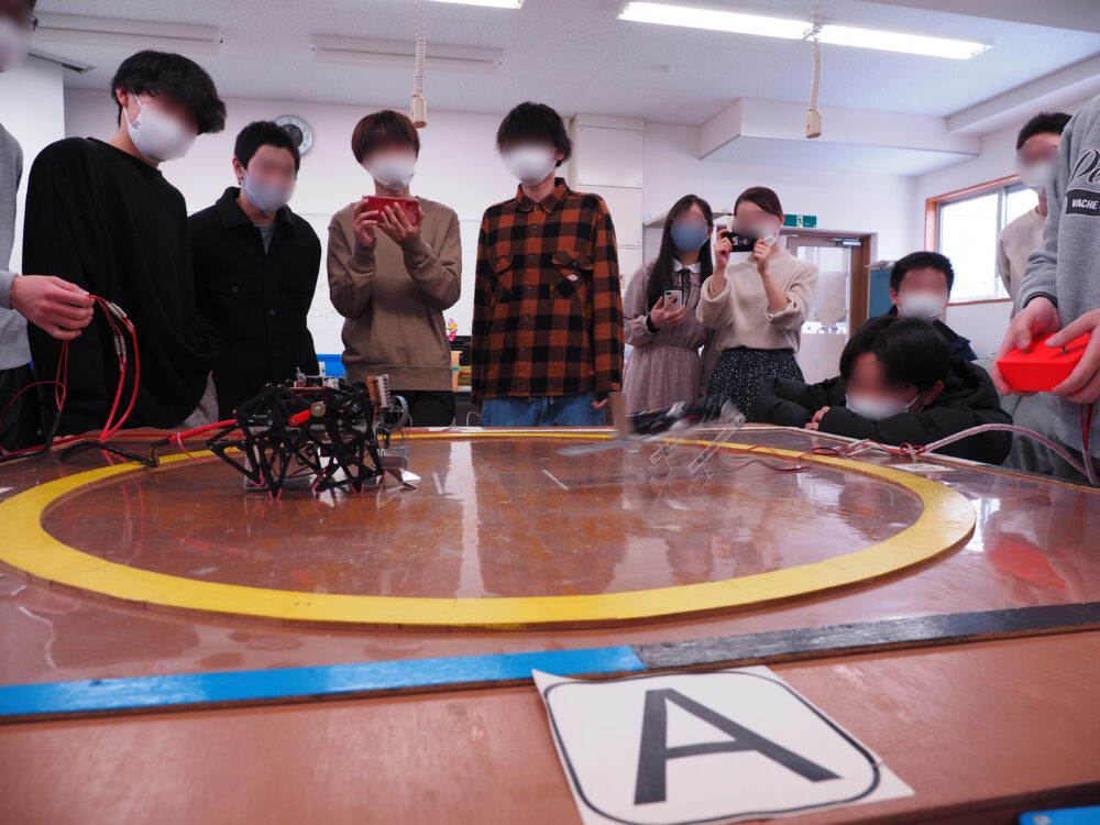 テーブル上の円の中で機械を動かす学生と、それを見守る学生たち。