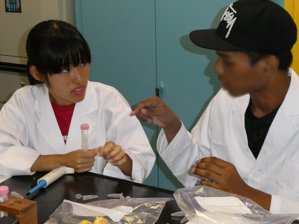 シンガポールからの留学生と、白衣を着て、実験器具を手に話し合っている学生の様子。