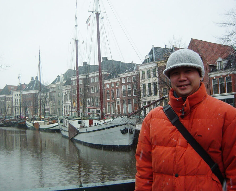 寒そうな白く曇ったなか、オランダらしい建物が並ぶ街並みを背景に、オレンジのダウンジャケットを着た笑顔の江崎先生