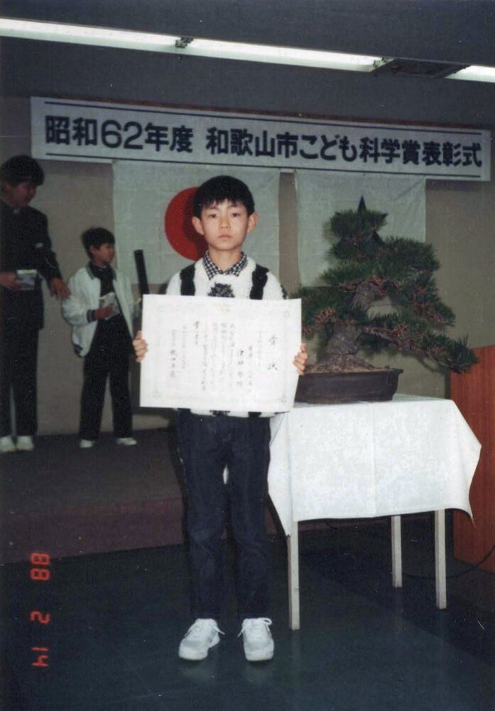 昭和62年度開催の和歌山市こども科学賞表彰式で、症状を持ち無表情の津田先生