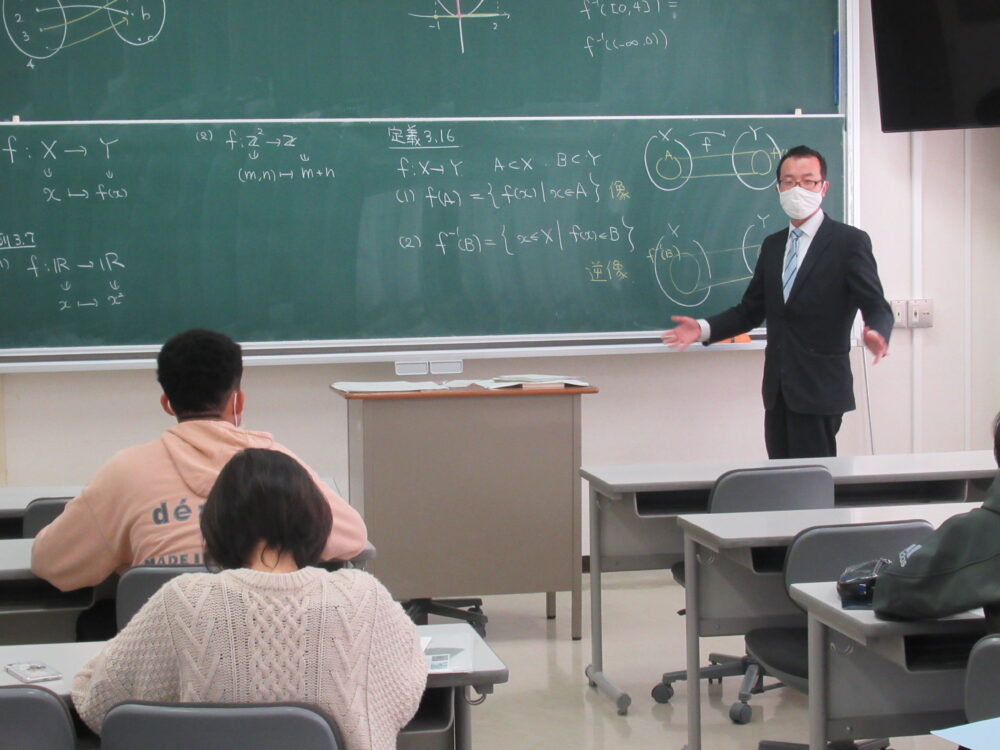 教壇で手振りを交えて学生に話す濵田先生