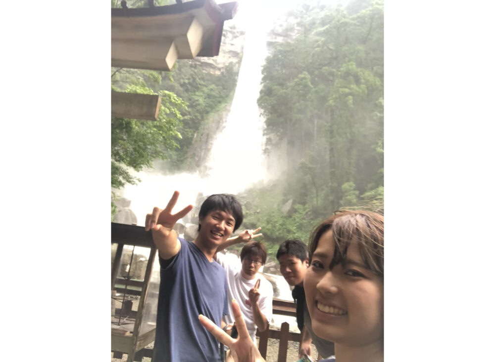 ゴーっと音が聞えてきそうな滝を背景にピースサインの嶋田さん。