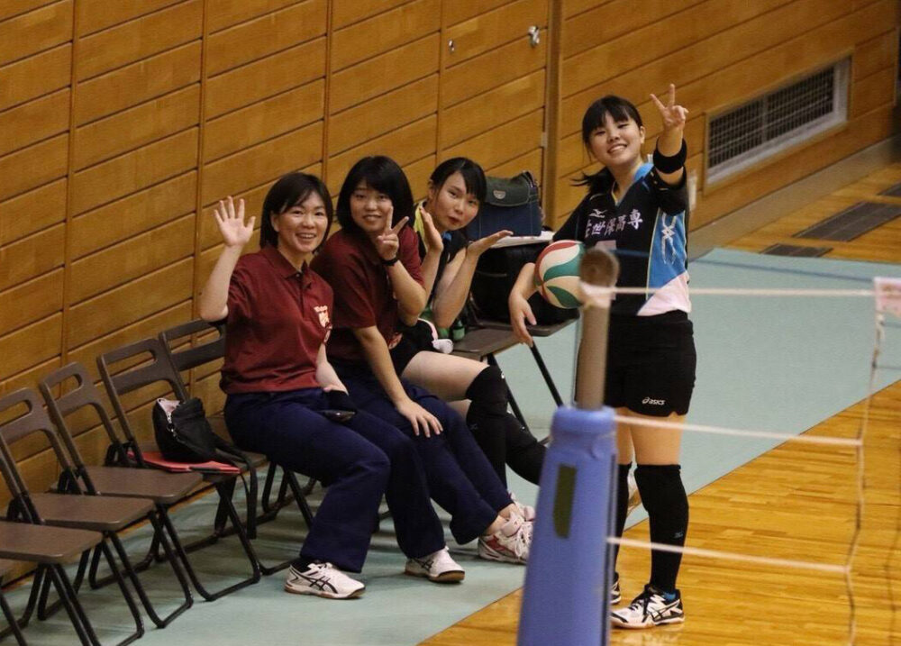 屋内のバレー場の椅子から、観覧席のカメラに向かって手を振る学生さんたちと大島先生