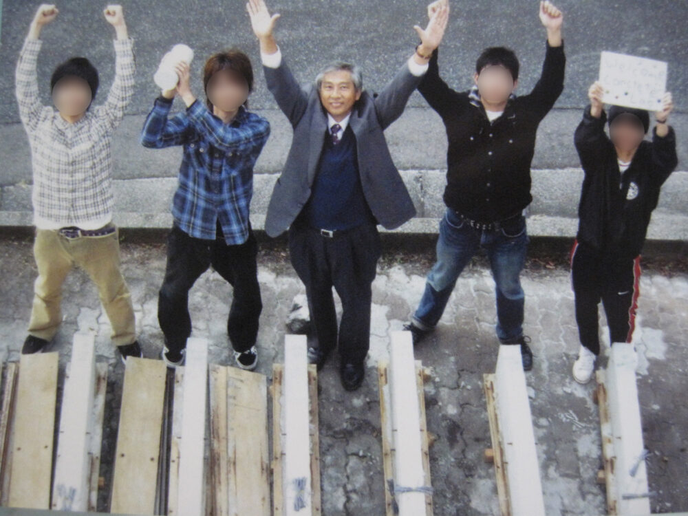 両サイドに2名ずつの男子学生さんとともに両手を挙げてわらう田村校長先生