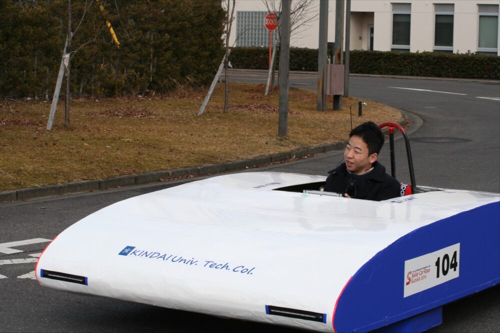 高専校内の道路で、未来的なかたちのソーラーカー（白色に、側面が青色）を操縦する木村さん