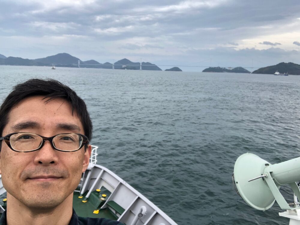 海上で、船の上で谷先生の自撮り。背景には山々と橋が。