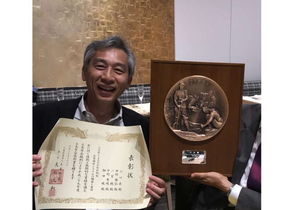 大きな銅の盾と賞状を手ににこやかな田村校長先生