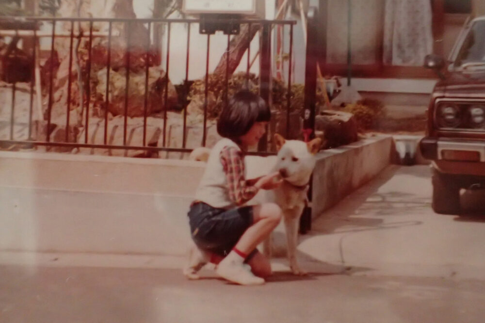 戸建ての家の前で、白の雑種犬と戯れる幼少期の磯村先生