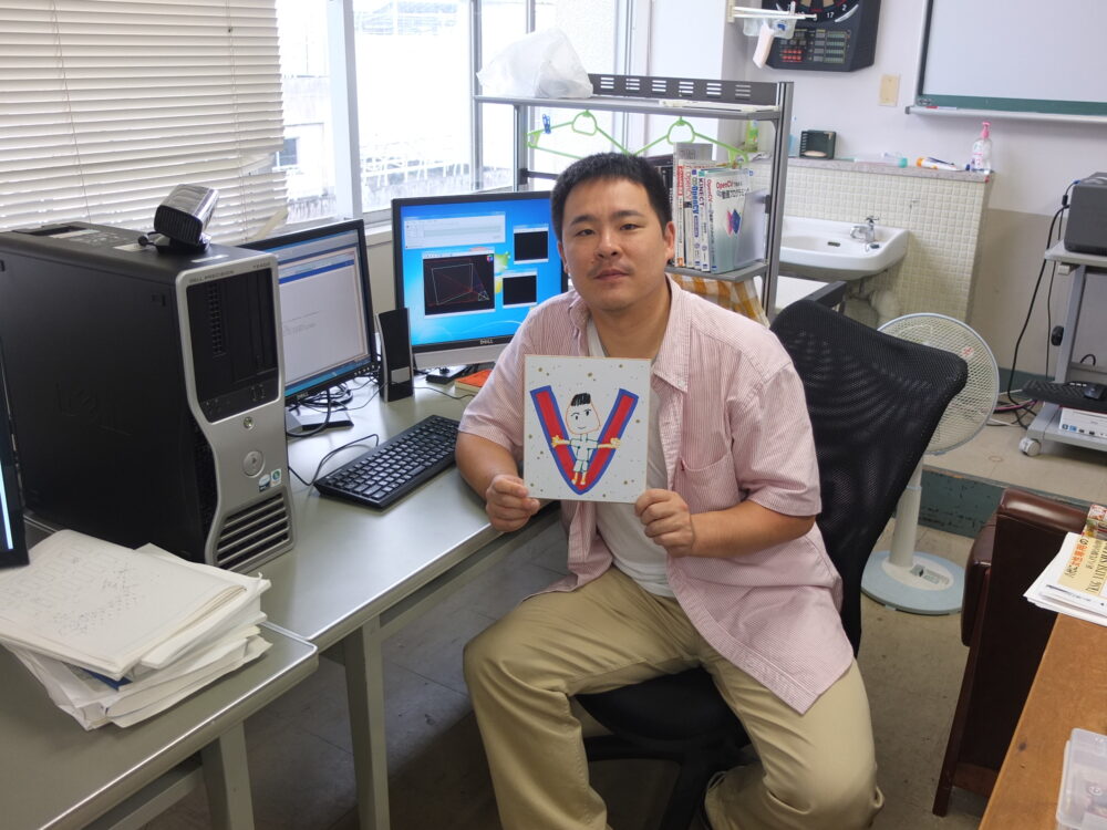 「V」という文字と寄せ書きがされた色紙を手に、デスクの椅子に掛けて、撮影