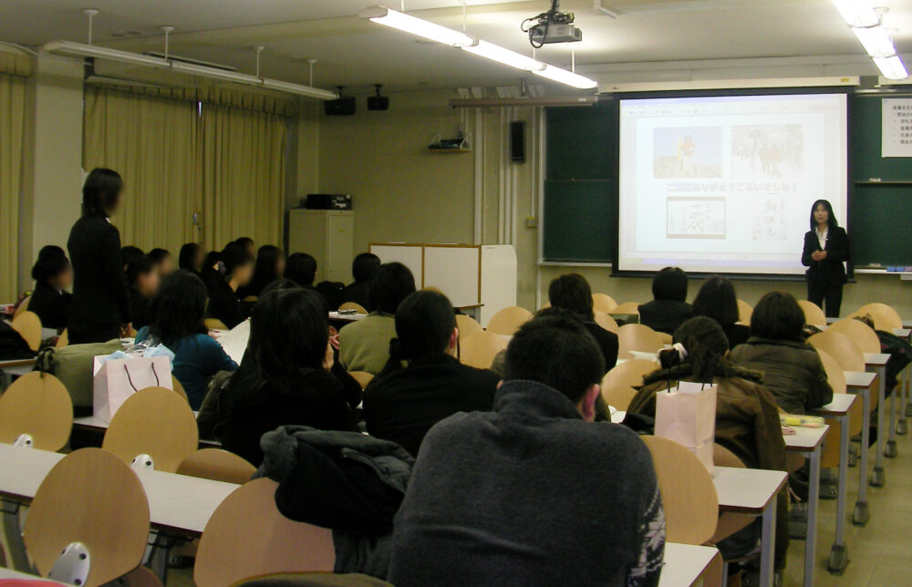 講義室で、多くの学生が着席するなか、前方の演台で、スクリーンにプロジェクターを投影し、講義を行う後藤校長