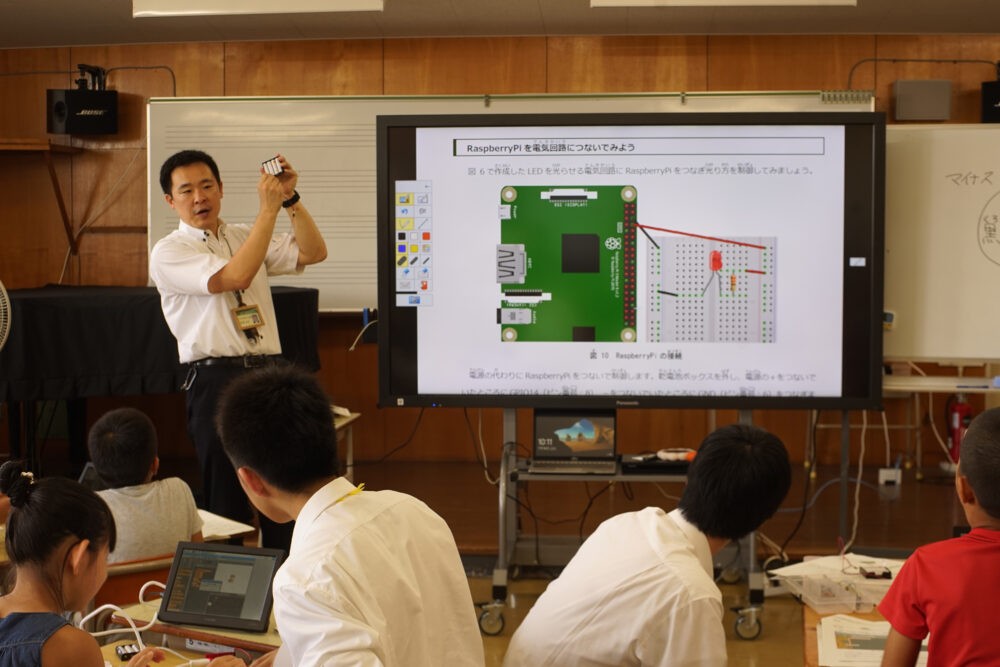 学生が小学生に寄り添って教えつつ、岩﨑先生は前方で全員に向けて説明を行っている