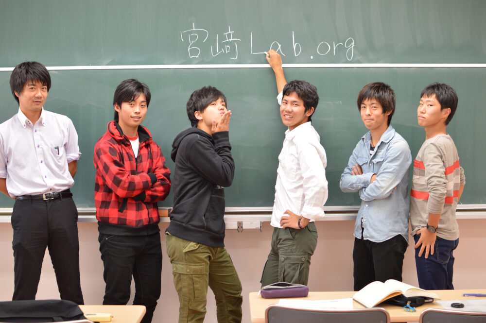 教室の緑の黒板に「宮﨑Lab」と書き、集合写真を撮る宮﨑研究室第1期生たち