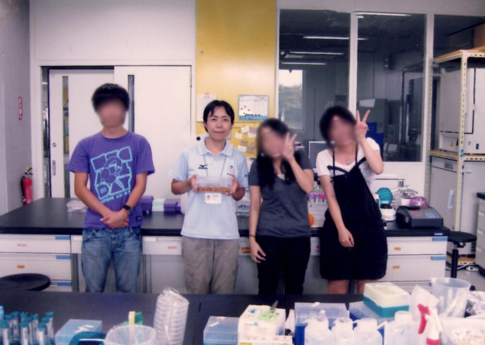 研究室で、先生の左に男子学生1名、右に女子学生2名とともに、集合写真