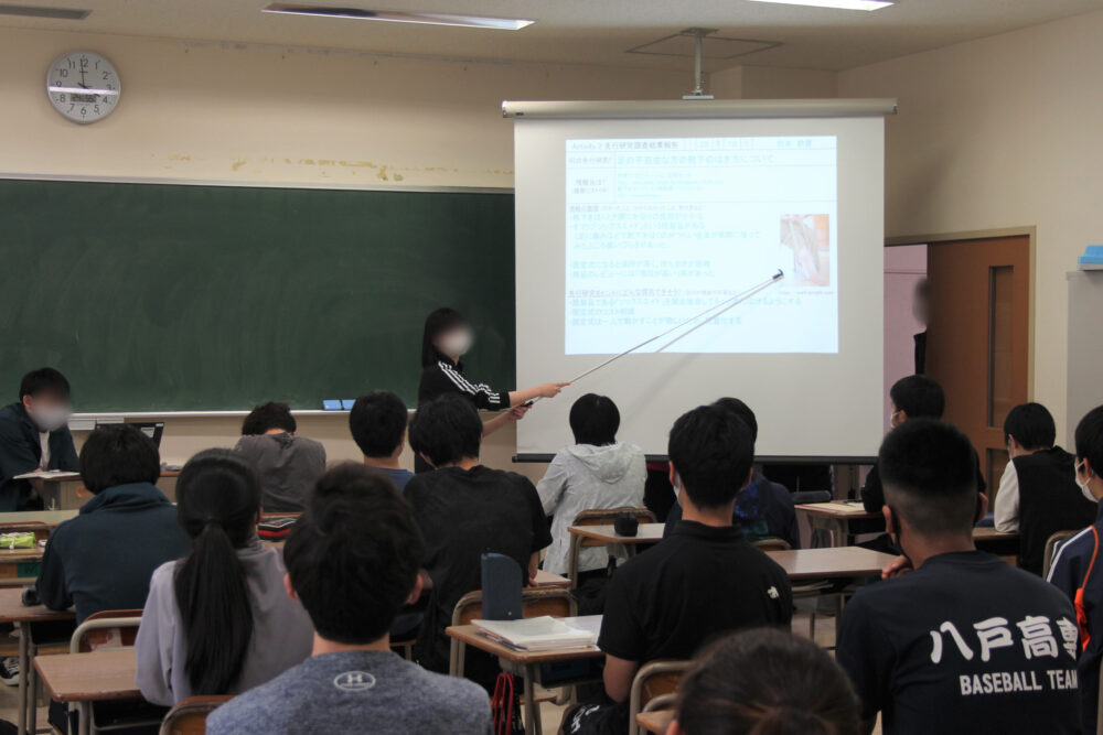 教室にて、前方の学生2名がスライドをプロジェクターで投影し、学生らに説明している様子