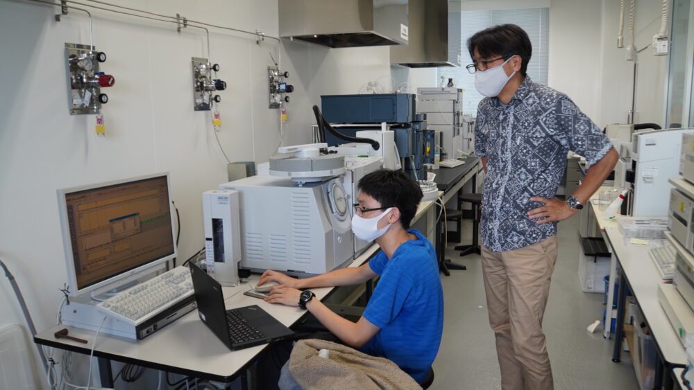 機材がおかれた研究室で、データを測定する学生に寄り添う玉城先生