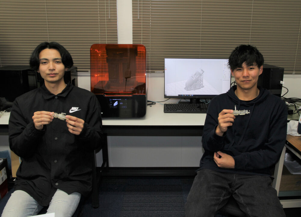研究室にて自らが設計し、3Ｄプリンターで製作した装置を手に持ってほほ笑む学生2名