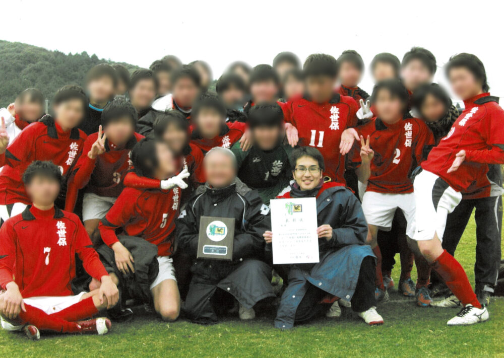 サッカー部の学生たちは、おそろいの白のパンツに赤のユニフォームを着て、楽しんでいる様子。そのなかに、賞状を持って映る松山先生