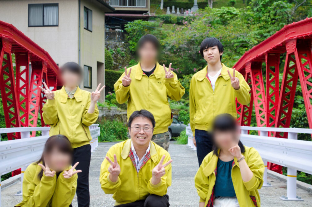 黄色のおそろいのジャケットを着て、赤い橋の上で集合写真をとる海田先生と山根さんたち