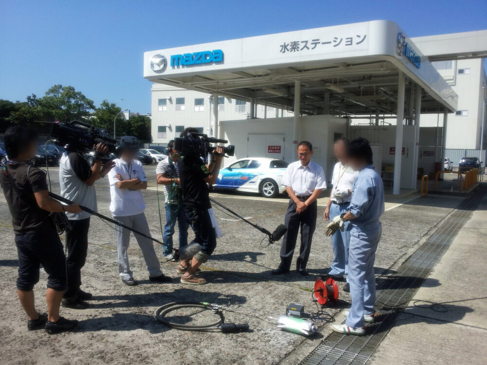 水素ステーションのあるスタンドで、大きなカメラやマイクに囲まれて取材を受ける野村先生と関係者たち