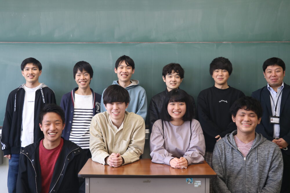 教室で、12期生9名とともに集合写真に写る青木先生