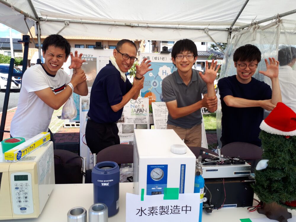 屋外のテントのもと、水素を使った実験を公開している野村先生と学生たち（同じポーズ）