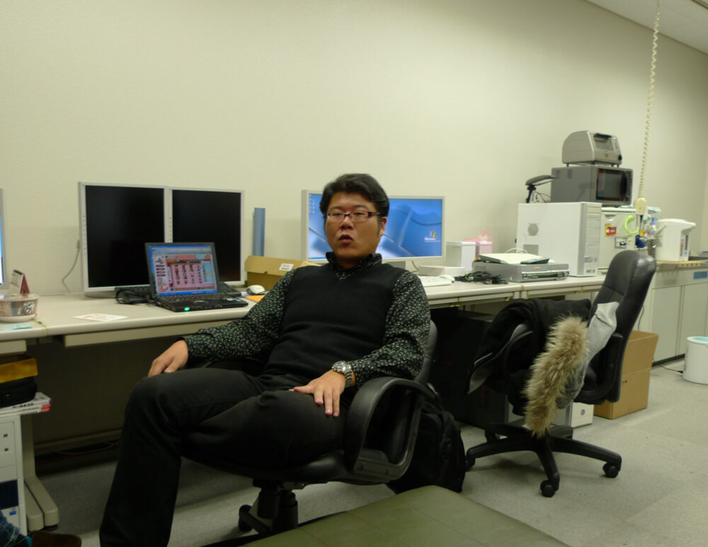 PCが並ぶデスクの椅子に座ってリラックスした感じの久保田先生