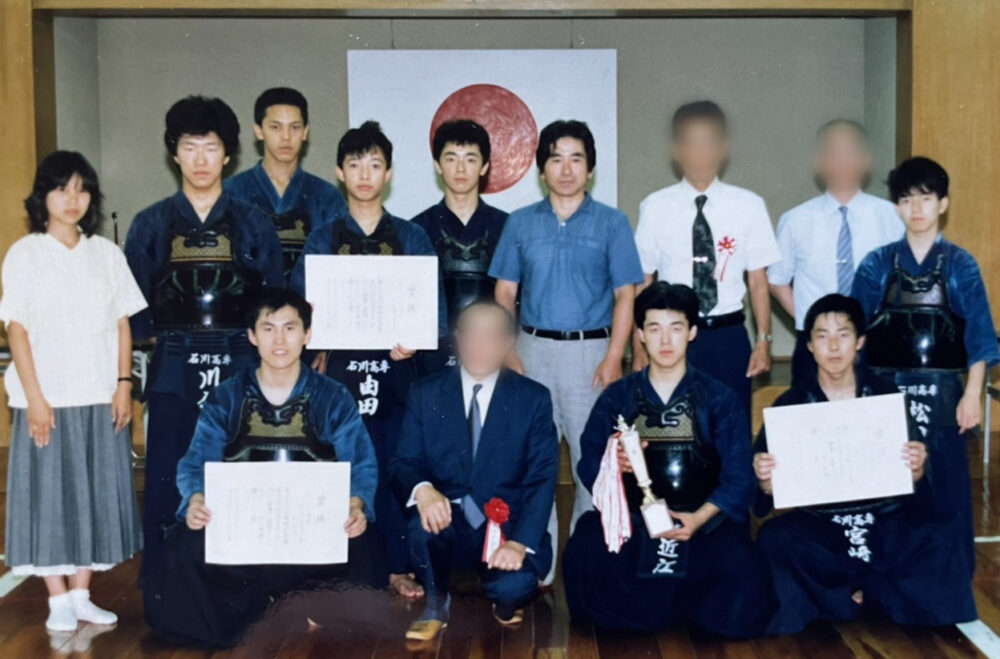 武道場にて、剣道着を身に付け、両手で賞状を持ち集合写真を撮る野村先生と仲間たち