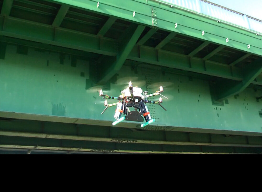 緑色の橋梁に向かって、6つのプロペラを回して飛ぶ機器