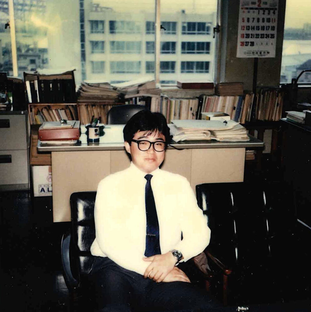 事務所内で、黒縁メガネをかけて、黒い応接セットに座りカメラに目線を送る清田先生