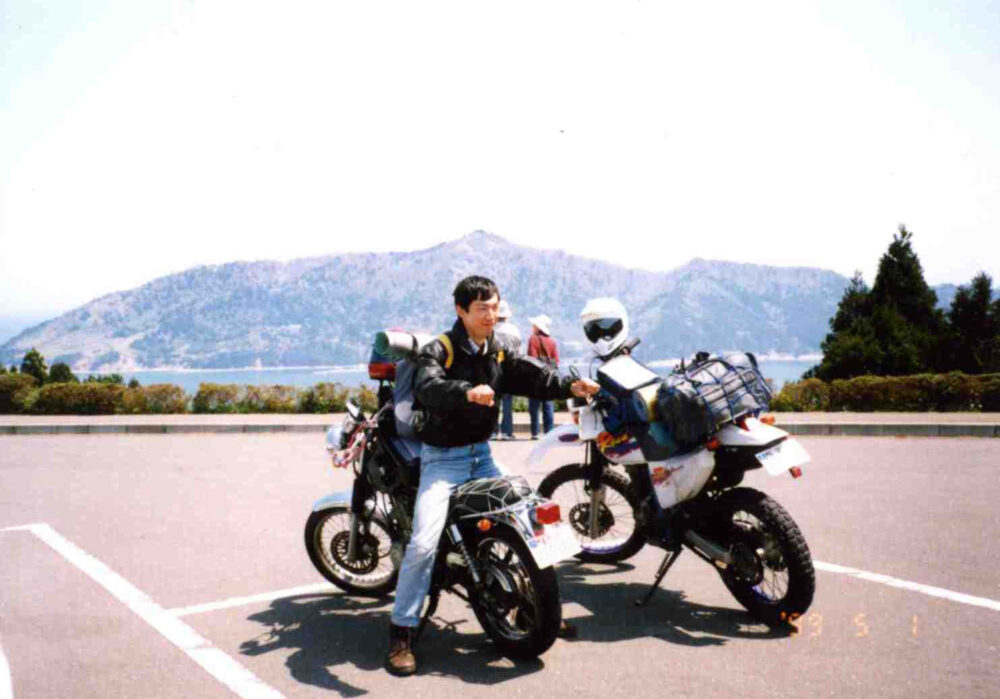 ツーリングで訪れた観光名所の駐車場で、バイクに反対向きにまたがり、ハンドルを握る仕草で写真におさまる平澤先生