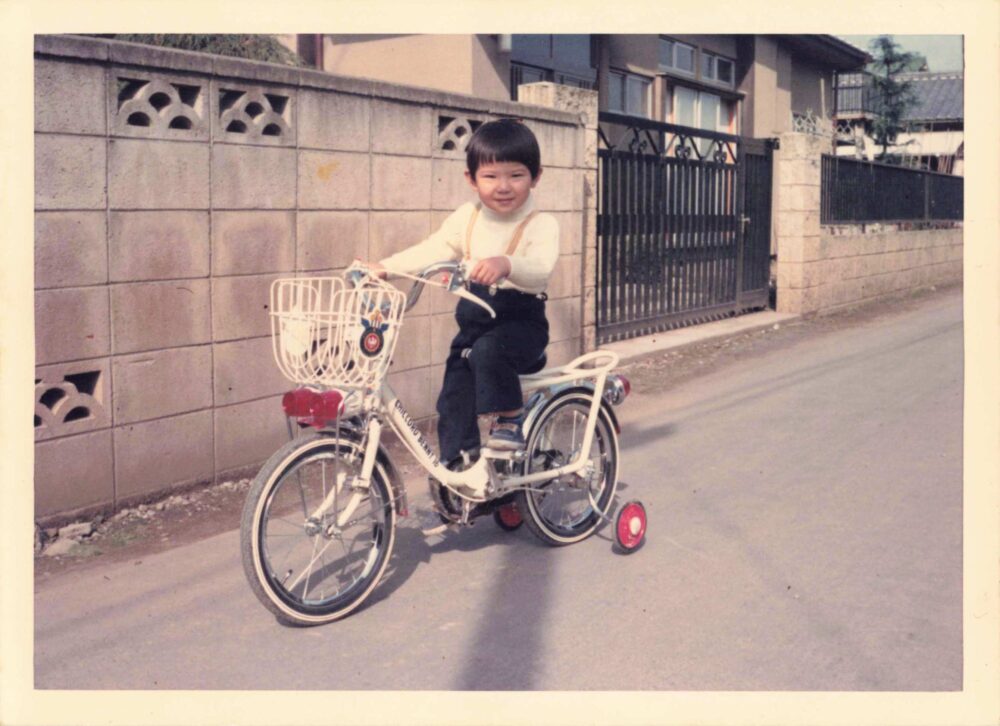 住宅街のブロック塀が続く家の前の道路で、補助輪付きの自転車をこぐ幼少期の村山先生
