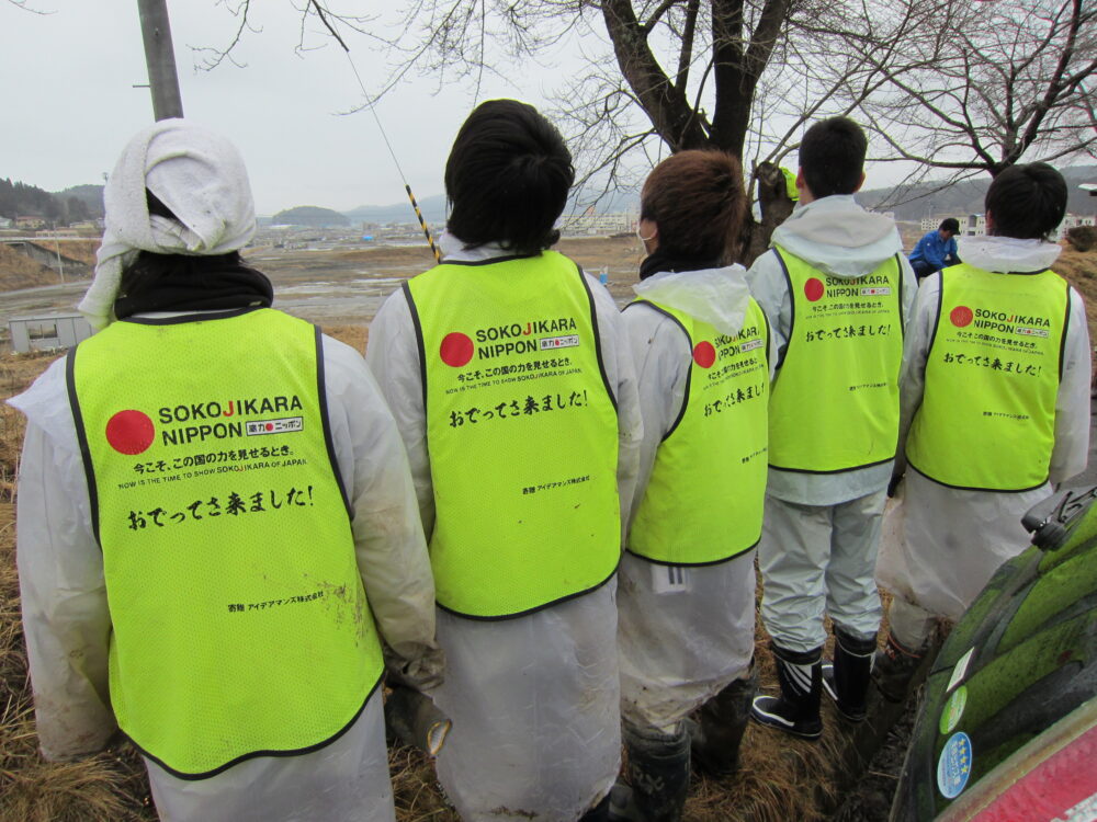 高低差のない、更地になった東日本にて、カッパに、蛍光黄色のビブスを身に付けボランティアをする学生たちの様子