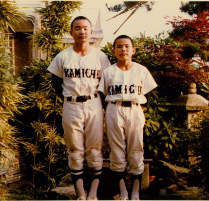 セピア色の写真。野球のユニフォームを身に付けた兄弟2人が家の前で記念撮影