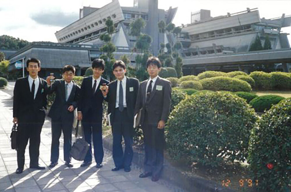 学会会場を背景に、スーツ姿の5名で集合写真