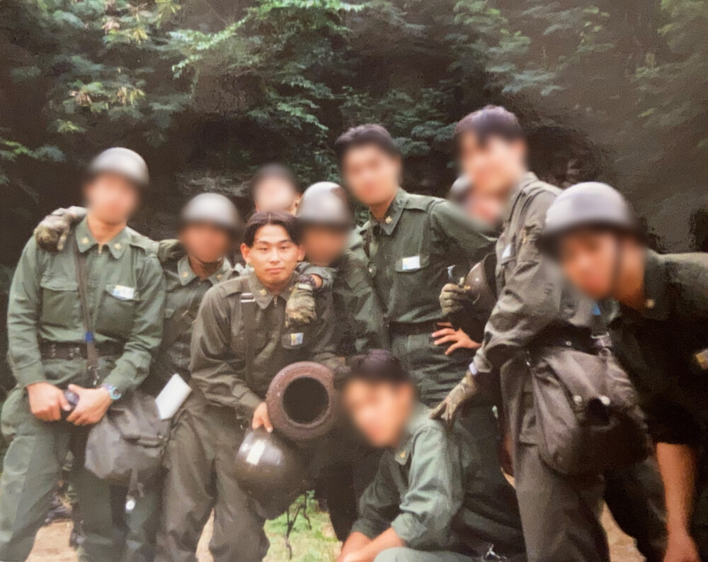 アーミー服に、濃茶のヘルメットをして、大砲を囲んで集合写真を撮る防衛大時代の村上先生