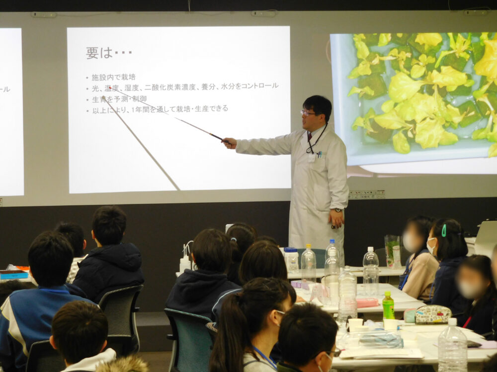 小学生たちが座る教室の前方で、大きなスクリーンにスライドを映し、白衣姿で説明する松浦先生