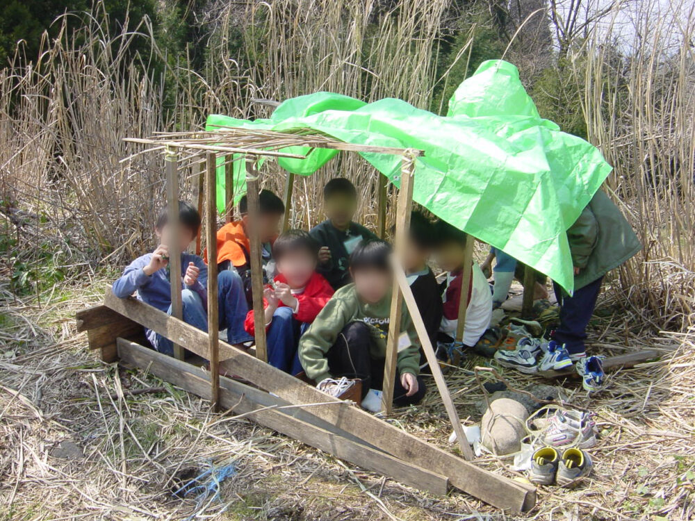 木の骨組みだけの簡素な小屋に、緑色のブルーシートを半分だけ掛けて、そのなかに子どもたちが座っている様子