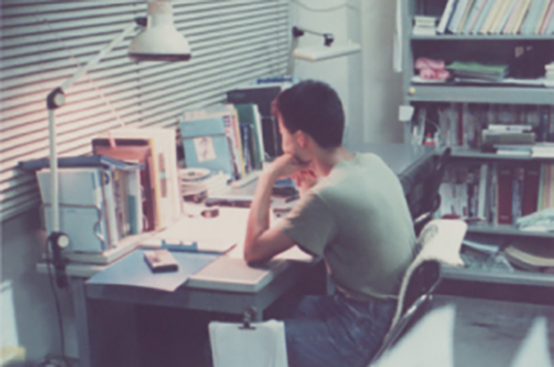 屋内、研究室の自席で、あごを左ひじに載せ、ノートを見つめる学生の頃の山田先生