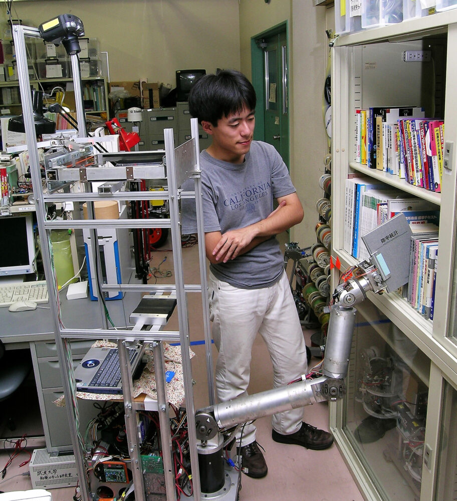 研究室にて、本棚から本を取り出すロボットとそれを見守る大学時代の冨沢先生