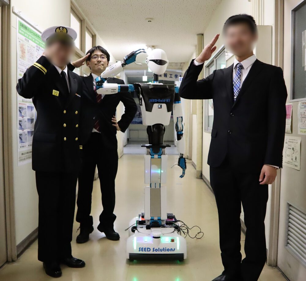 大学内の廊下で、上半身がヒト型のロボットが敬礼している左右に、人物も敬礼している