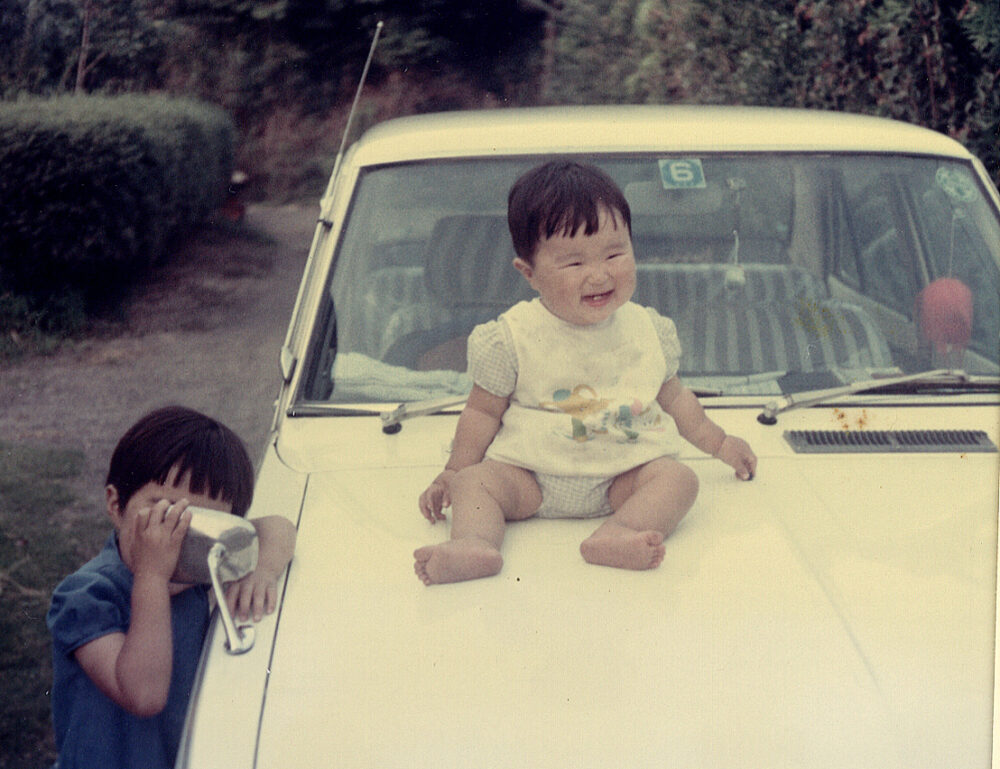 前方にカーブミラーが付いている型のクリーム色の自動車のボンネットの上に座る、幼少期の徳楽先生