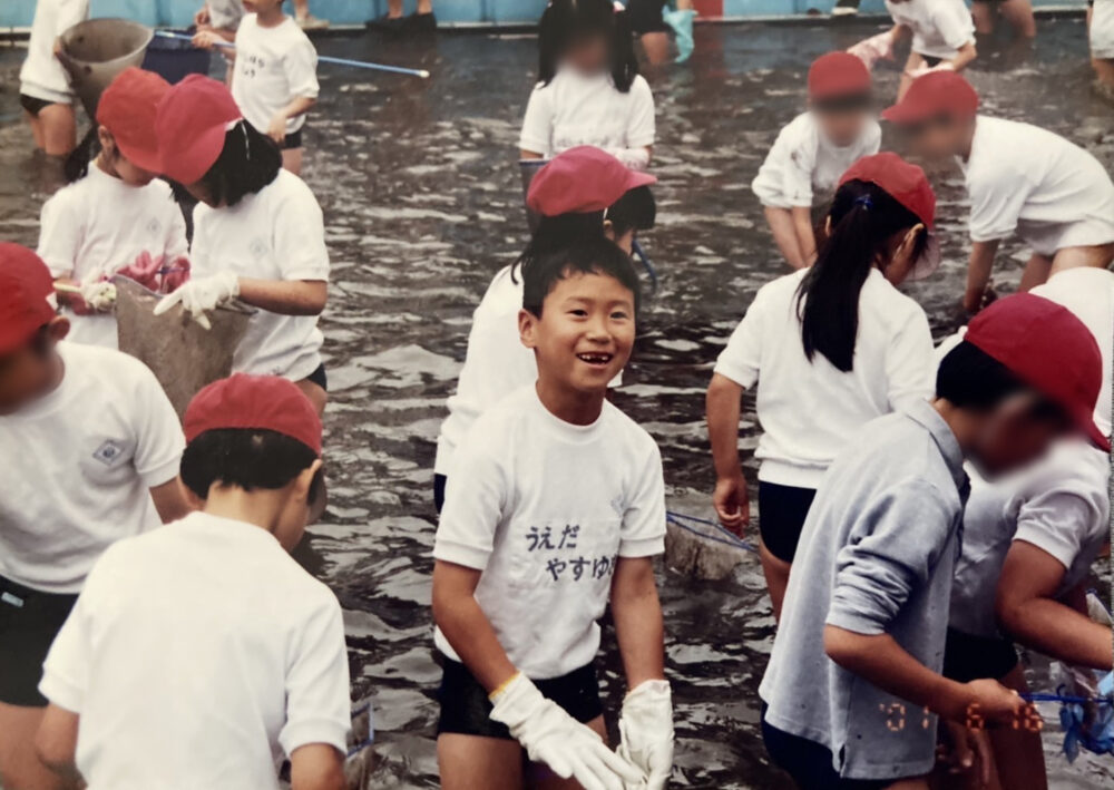 みな体操着姿で、同級生たちと、膝くらいまで水に浸かって何かを採集しているなか、中央でこちらに笑顔を向ける少年時代の植田先生